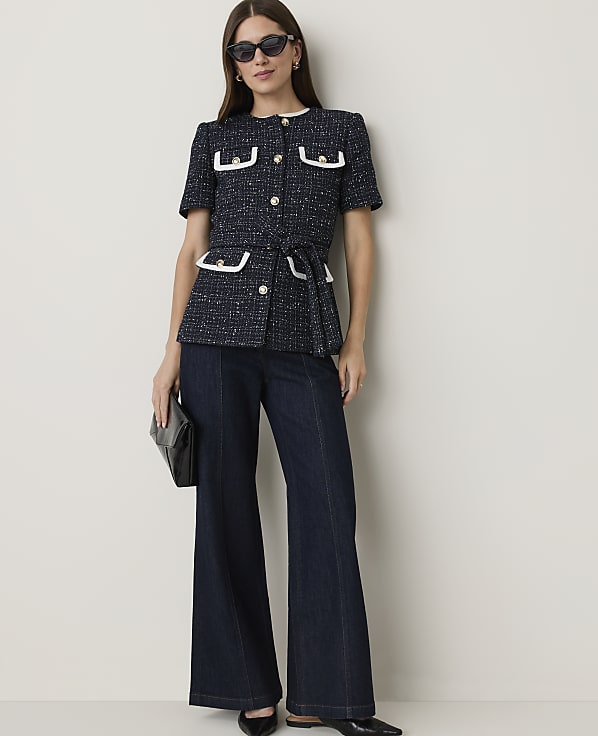 A woman in a navy tweed jacket and dark flared jeans stands against a plain, light-colored wall, holding a black clutch.