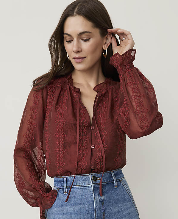 A woman in a rust-colored lace blouse and blue jeans poses against a plain, light background.