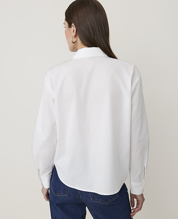 A woman in a white button-down shirt and blue jeans stands against a plain white background.