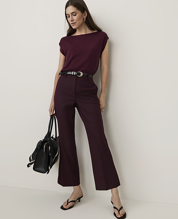 A woman in a burgundy top and matching flared pants stands against a plain white background, holding a black tote bag.