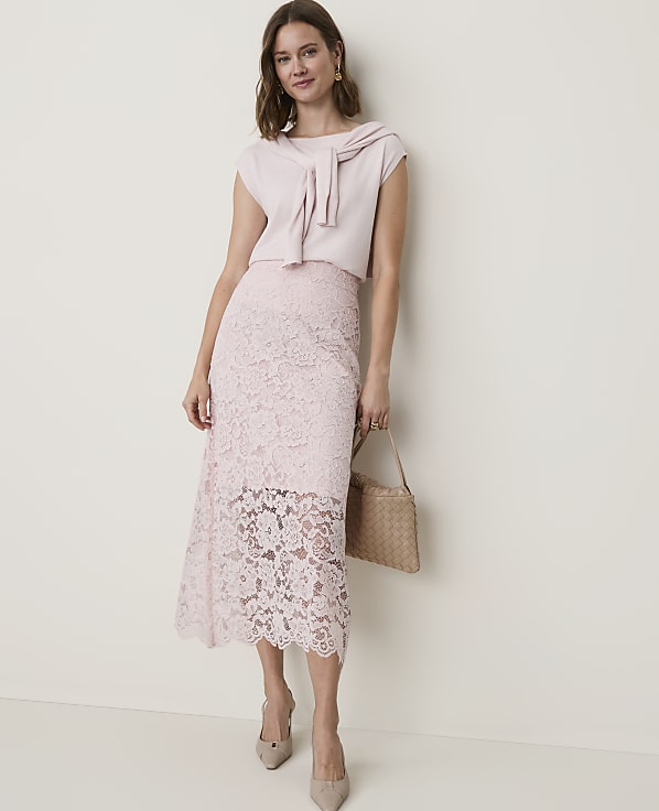 A woman in a light pink lace skirt and matching top stands against a plain, light-colored wall, holding a woven handbag.