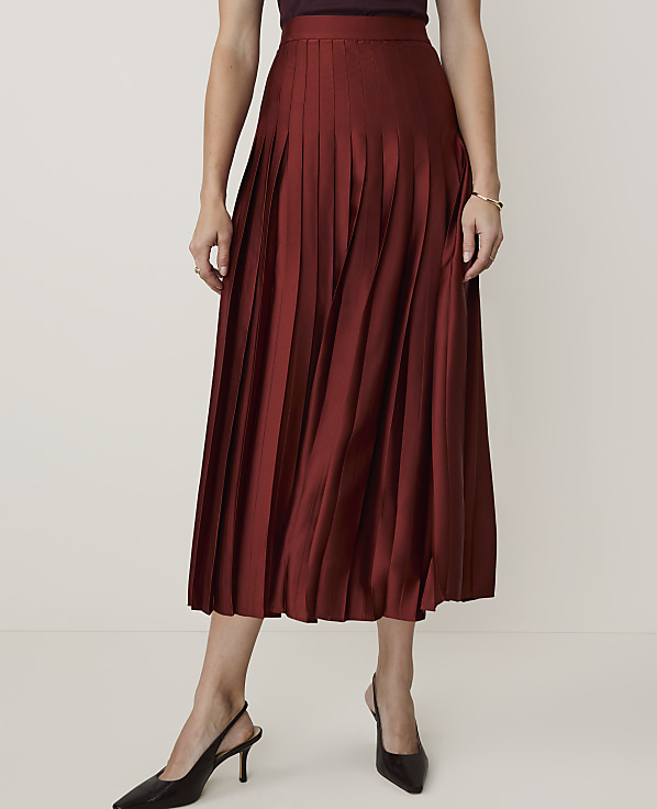 A woman in a rust-colored pleated midi skirt and black slingback heels stands against a plain, light-colored background.