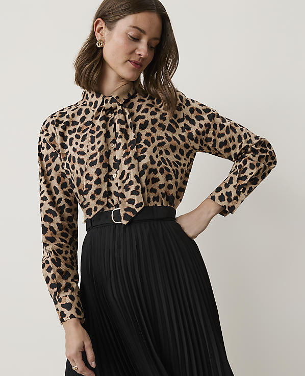 A woman in a leopard print blouse and black pleated skirt stands against a plain, light-colored background.