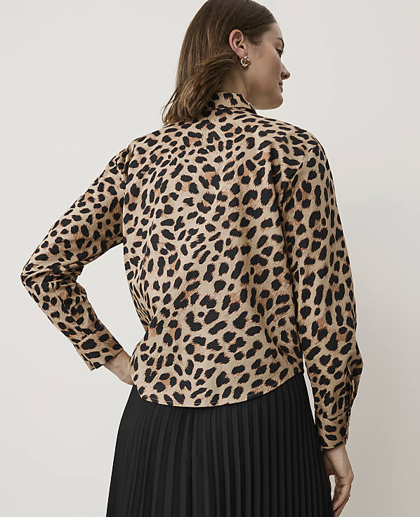 A woman in a leopard print shirt and black pleated skirt stands against a plain background.