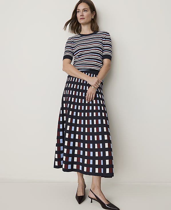 A woman in a striped top and a patterned skirt stands against a plain wall.