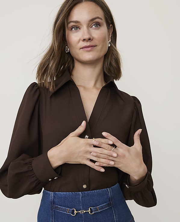 A woman in a brown blouse and blue skirt poses with her hands near her chest against a plain background.