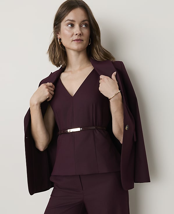 A woman in a burgundy suit stands against a plain, light-colored background, holding the lapels of her jacket.