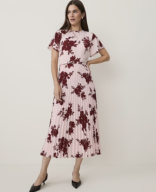 A woman in a pink floral print top and pleated skirt stands against a plain, light background.