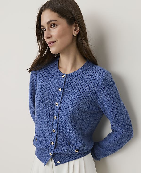 A woman in a blue textured cardigan with pearl buttons stands against a plain, light-colored background.