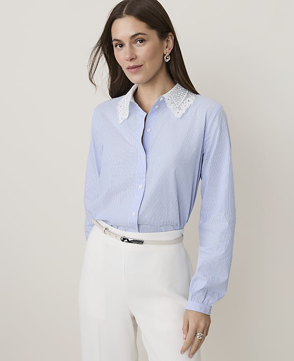 A woman in a blue pinstripe shirt with a lace collar and white pants stands against a plain, light background.