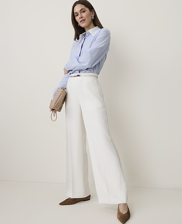 A woman in a blue striped shirt and white wide-leg pants stands against a plain, light-colored background, holding a small woven purse.