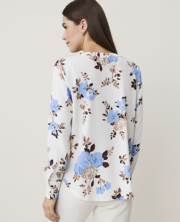A woman in a white floral blouse stands against a plain, light background.
