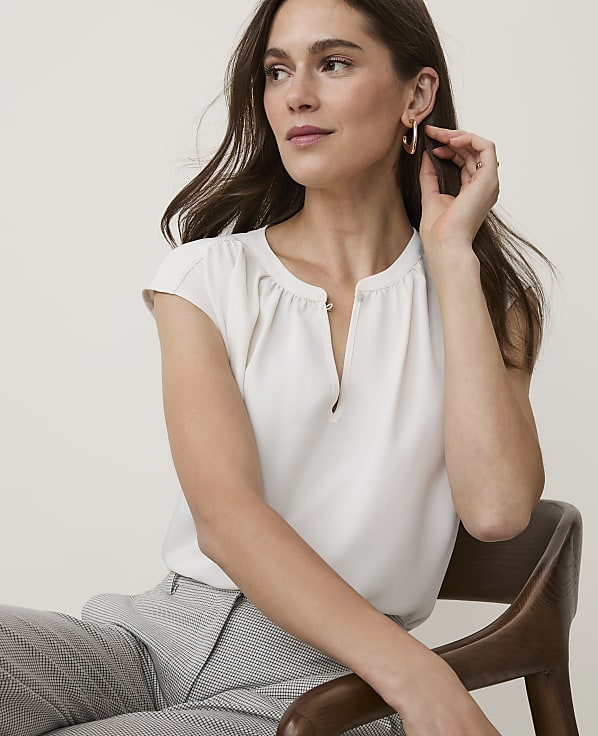 A woman in a cream blouse and patterned pants sits on a wooden chair against a plain background.