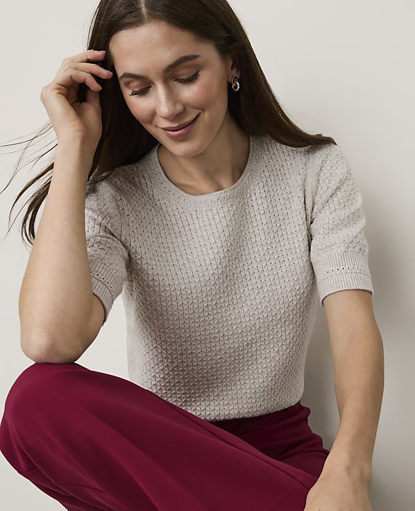 A woman in a light-colored textured sweater and maroon pants smiles softly against a plain background.