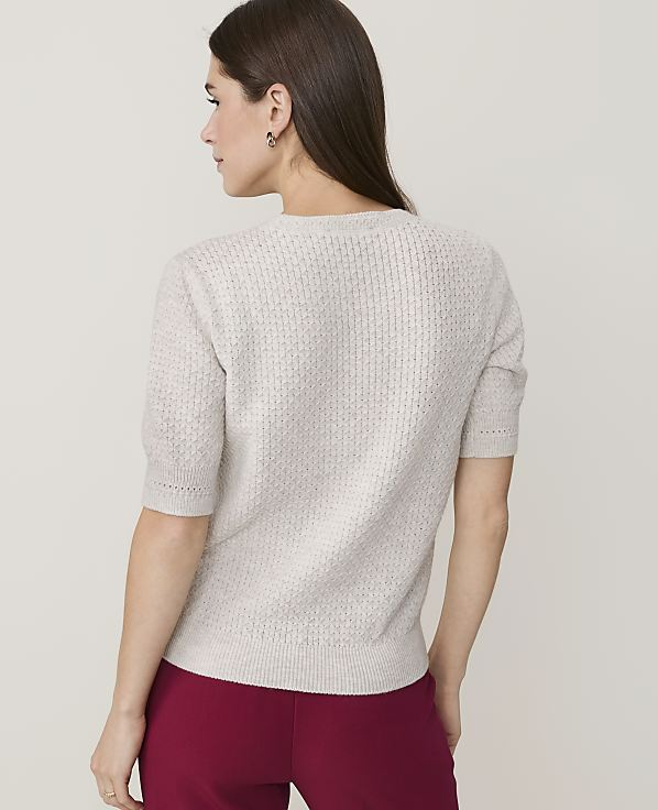 A woman in a light-colored, textured sweater and red pants stands against a plain, light background.