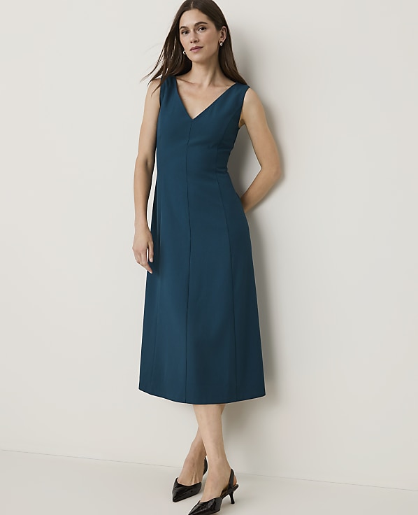 A woman in a teal V-neck dress and black heels stands against a plain, light-colored wall.