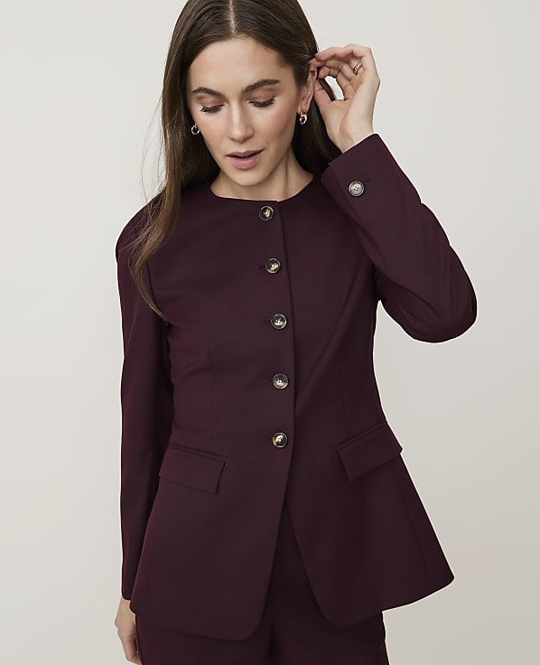 A woman in a burgundy blazer with round buttons adjusts her earring against a plain white background.
