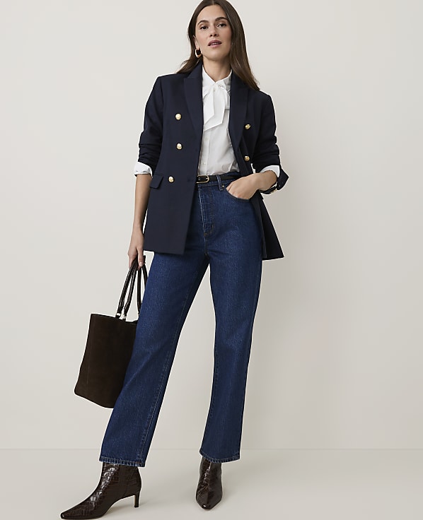 A woman in a navy blazer and jeans stands against a plain background, holding a dark tote bag.