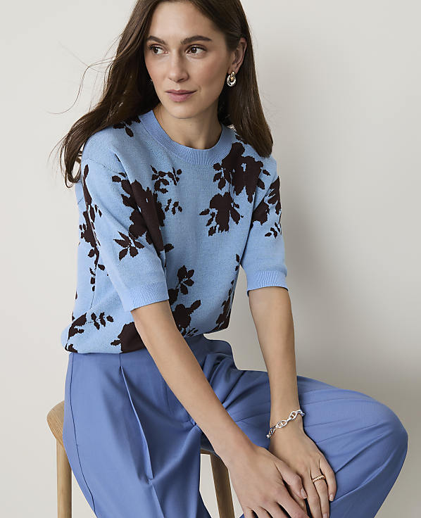 A woman in a light blue floral sweater and matching blue pants sits against a plain, light background.