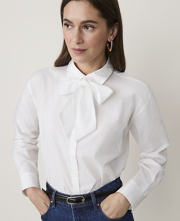 A woman in a white bow-neck blouse and blue jeans stands against a plain, light background.