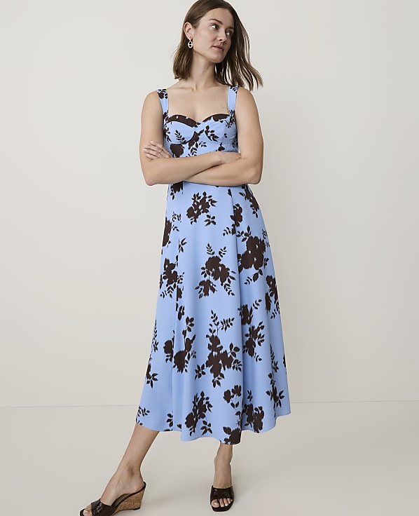A woman in a blue floral dress stands with her arms crossed against a plain, light-colored background.