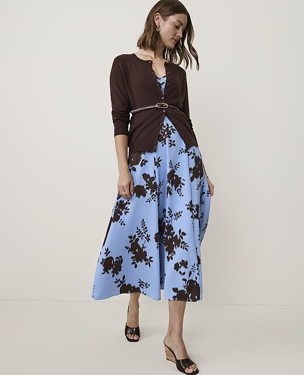 A woman in a brown cardigan and a blue floral dress stands against a plain, light-colored background.