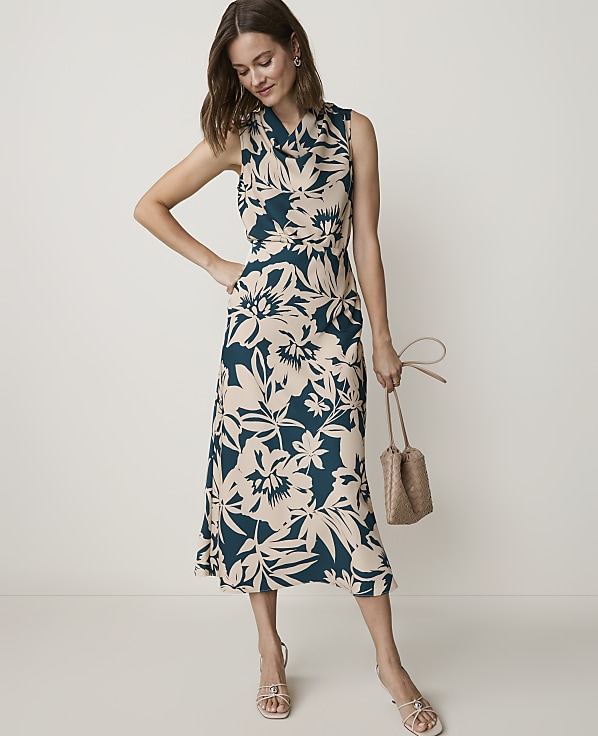 A woman in a floral dress and sandals stands with a woven bag against a plain background.