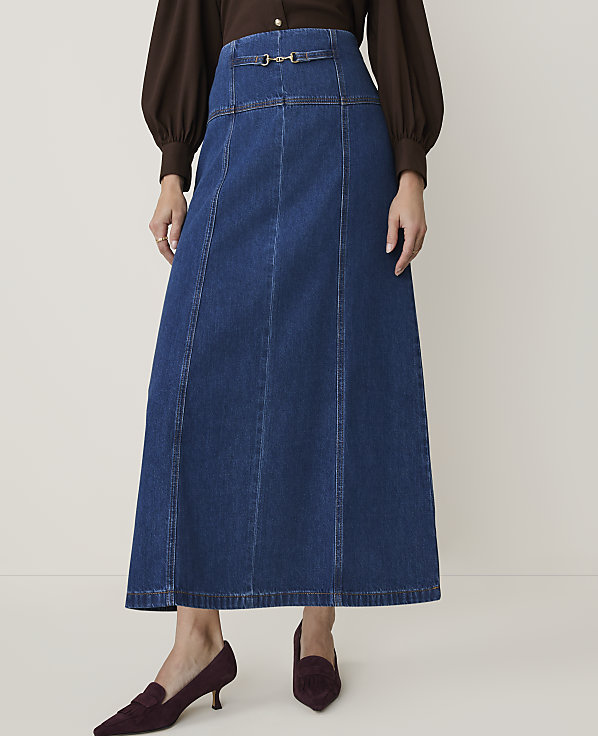 A woman in a brown blouse and a dark denim maxi skirt with a buckle detail stands on a light-colored floor, wearing burgundy loafers.