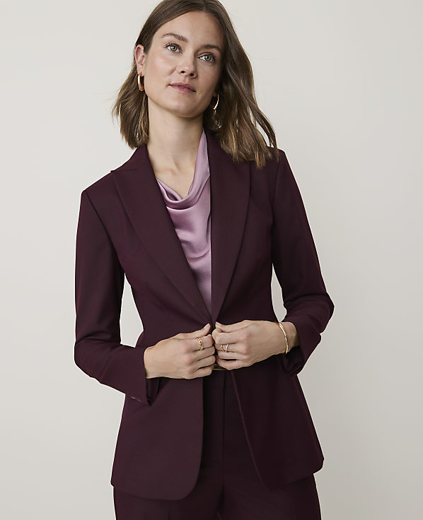 A woman in a burgundy blazer and pink blouse stands against a plain, light-colored background.