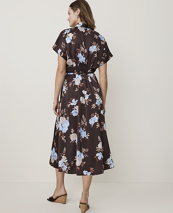 A woman in a brown floral dress stands against a plain, light-colored background.