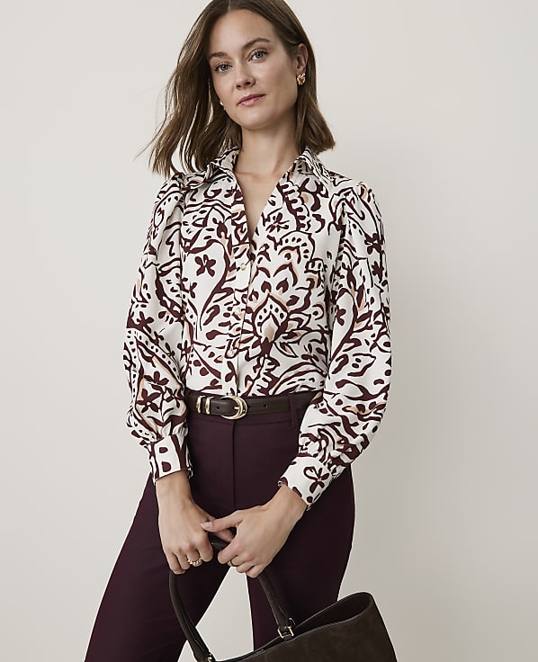 A woman in a patterned blouse and burgundy pants holds a dark brown bag in front of a plain, light-colored background.