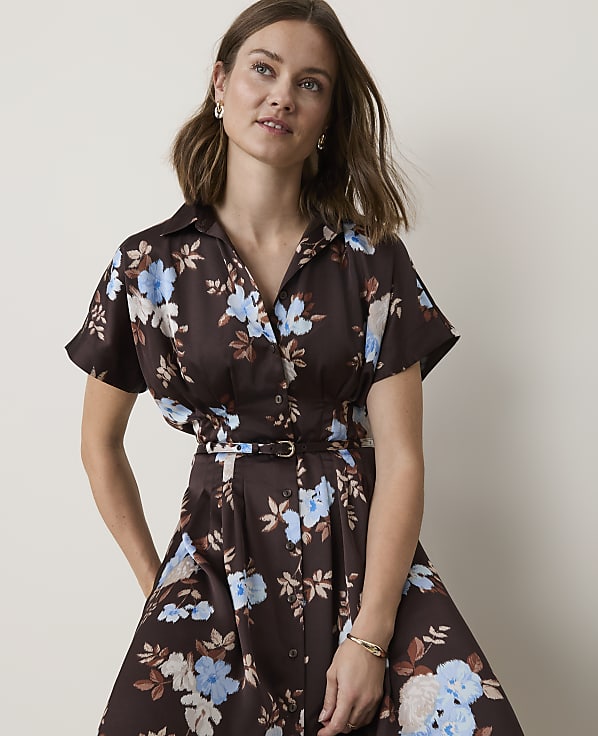 A woman in a brown floral dress with a cinched waist stands against a plain white background.