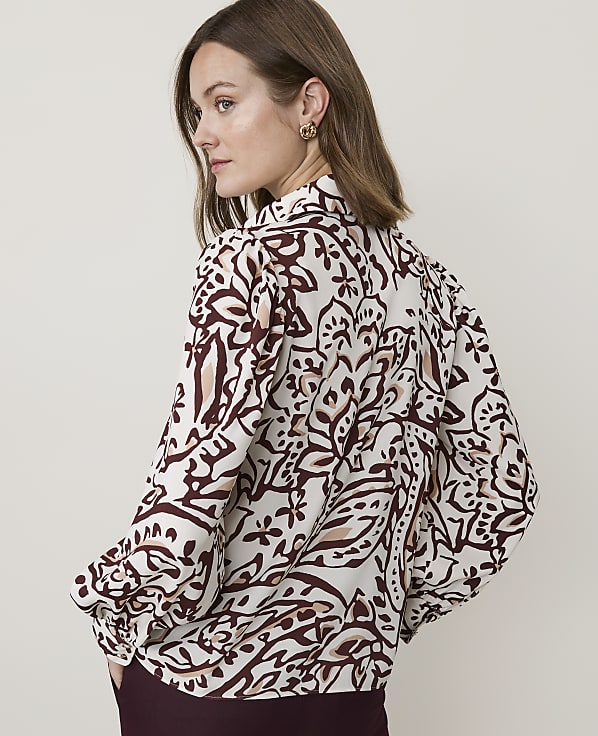 A woman in a patterned blouse stands against a plain, light background.