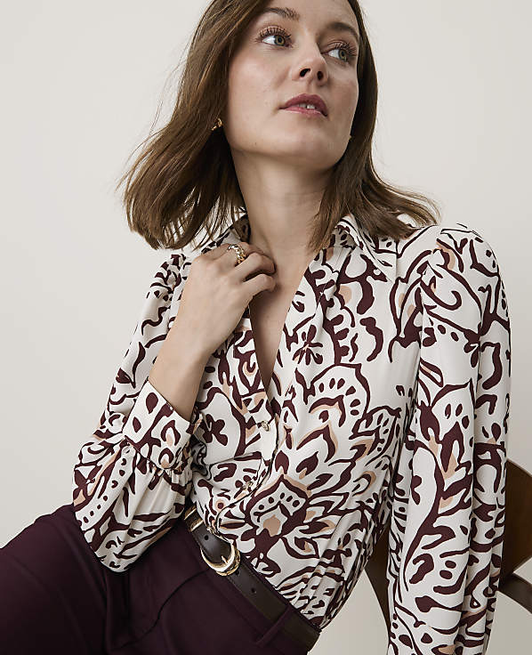 A woman in a patterned blouse and burgundy pants is seated against a plain background.