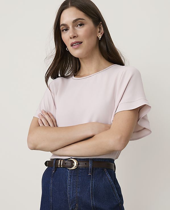 A woman in a pink blouse and denim jeans stands with her arms crossed against a plain background.