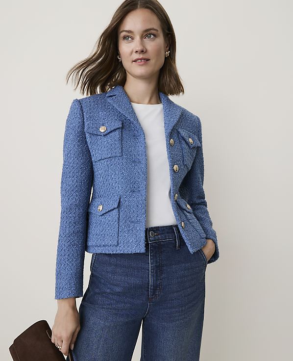 A woman in a blue tweed jacket and jeans stands against a plain, light background, holding a brown clutch.