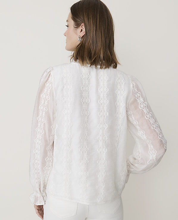 A woman in a white lace-trimmed blouse stands against a plain, light-colored background.