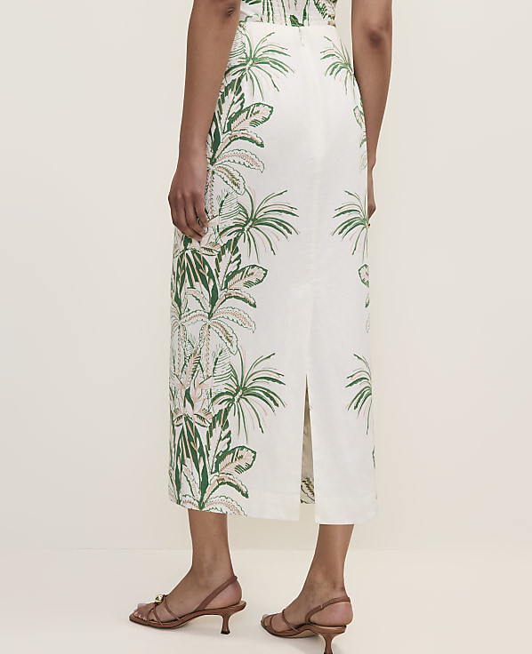 A person stands in the foreground wearing a white skirt with a green tropical leaf pattern, while the background is a plain, light-colored wall.