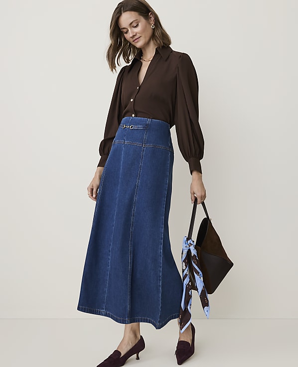 A woman in a brown blouse and denim skirt stands against a plain, light-colored background, holding a dark handbag with a scarf.