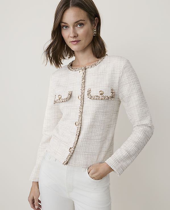 A woman in a cream tweed jacket and white pants stands against a plain, light background.