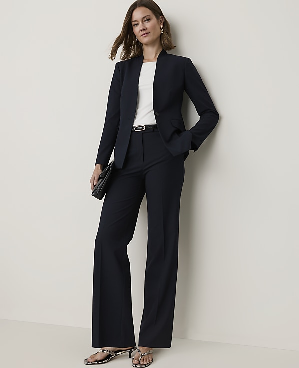 A woman in a navy pantsuit stands against a plain, light-colored wall, holding a black clutch.