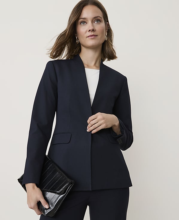 A woman in a navy blazer and pants holds a black clutch in front of a plain background.