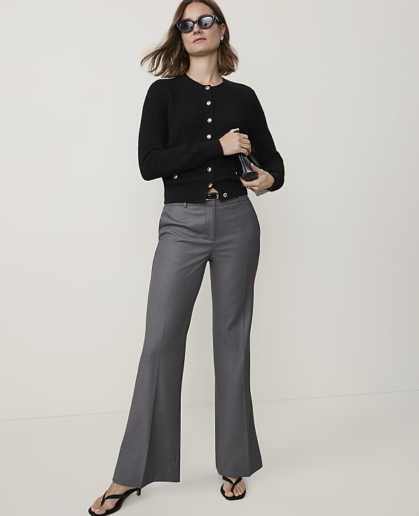 A woman in a black cardigan and gray flared pants stands against a plain, light-colored background.