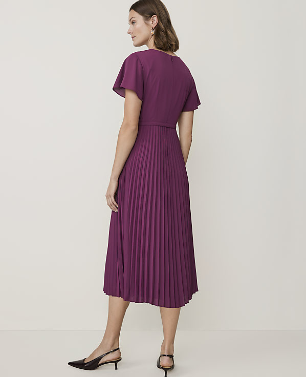 A woman in a purple pleated dress stands against a plain white background.