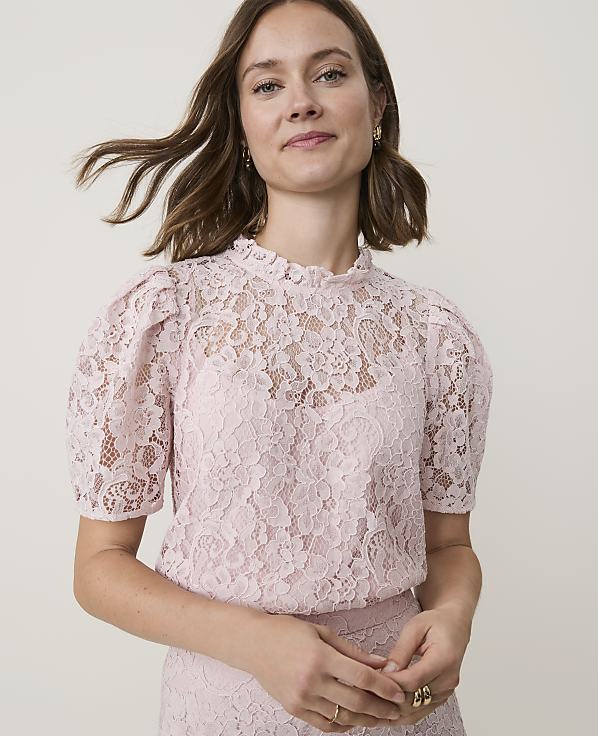 A woman in a pink lace top with puffed sleeves stands against a plain, light background.