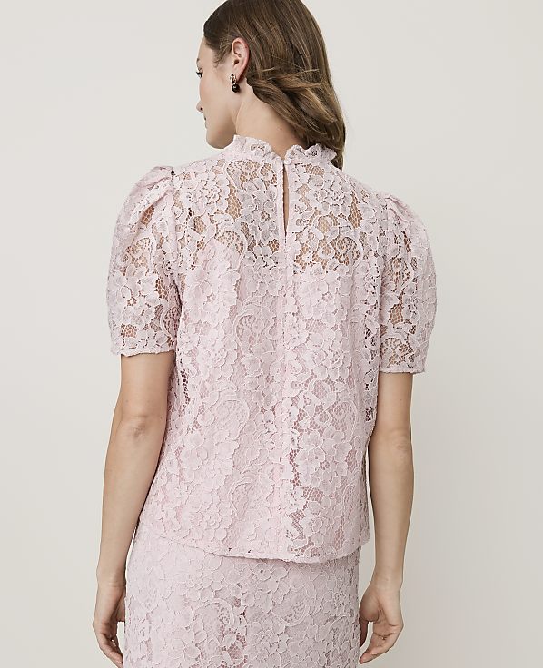 A woman in a pink lace top with puffed sleeves stands against a plain white background.