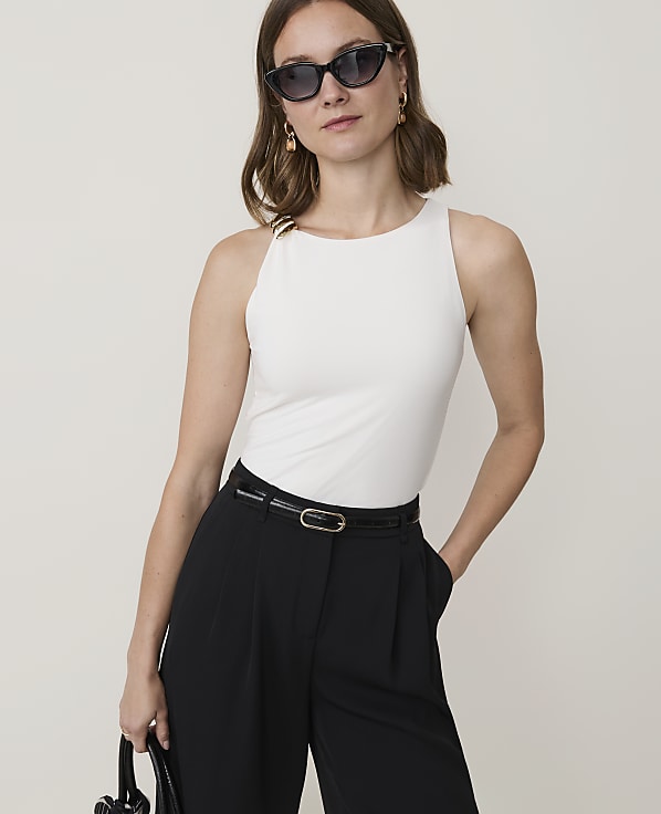 A woman in a white sleeveless top and black pants stands against a plain background.