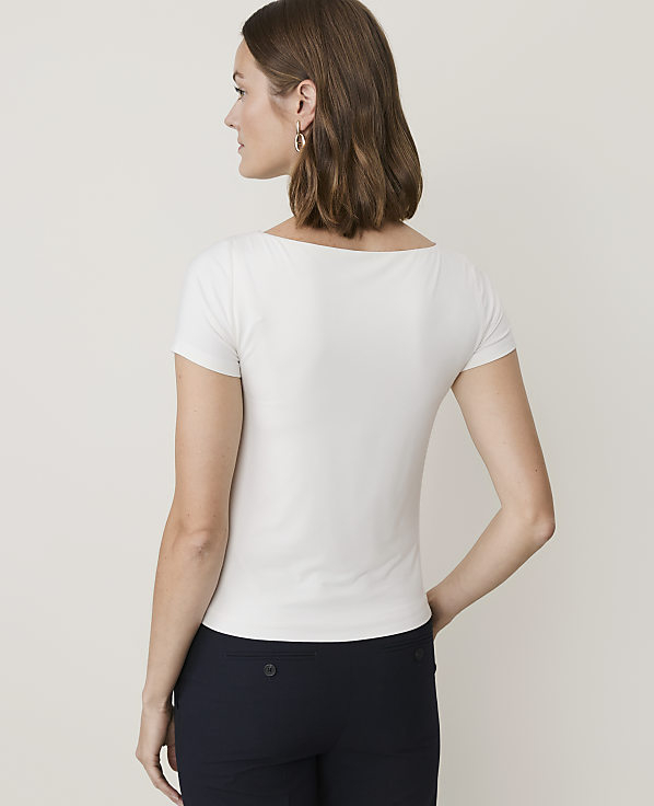 A woman in a white top and dark pants stands against a plain, light background.