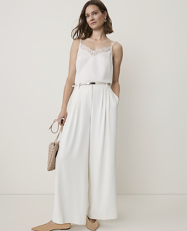A woman in a white camisole and wide-leg pants stands against a plain, light background, holding a woven purse.