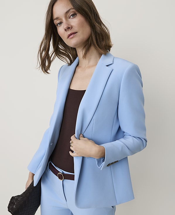 A woman in a light blue blazer and matching pants stands against a plain, light background.